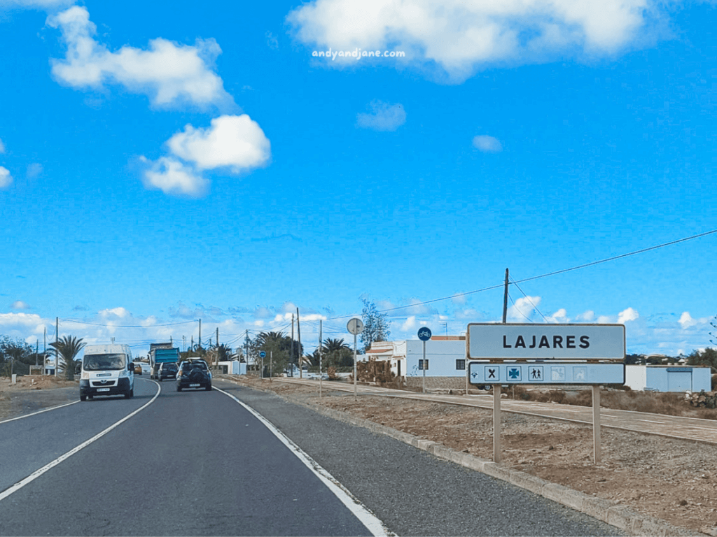 A road sign indicating "Lajares," surrounded by a clear blue sky, with vehicles on a winding road and buildings in the background.