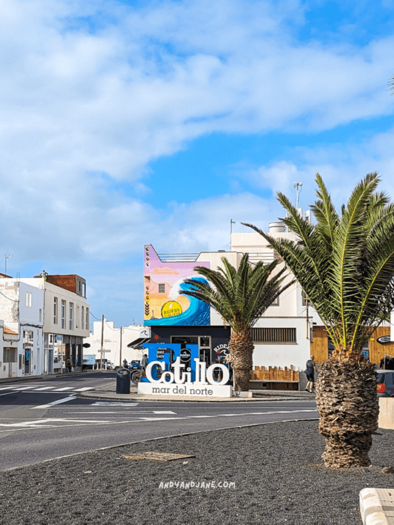 A bustling street view in El Cotillo featuring palm trees, a vibrant mural, and a sign reading "cotillo mar del norte."