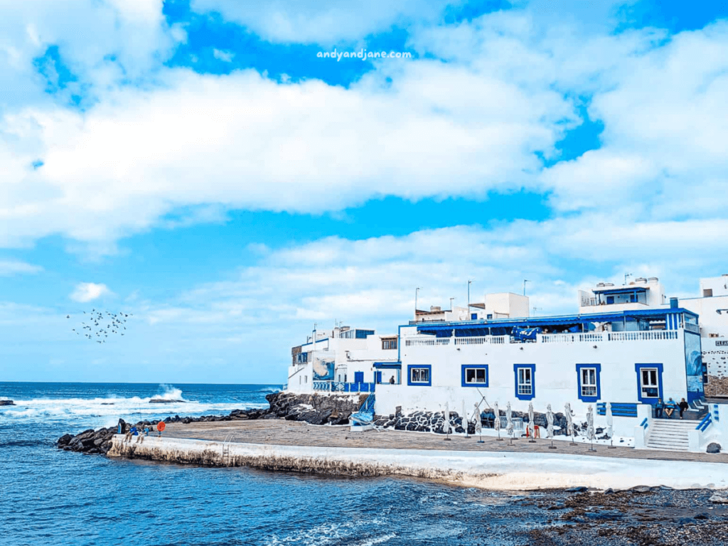 Coastal scene in El Cotillo with white buildings by the sea, rocky shore, and blue skies dotted with clouds, evoking a tranquil seaside atmosphere.