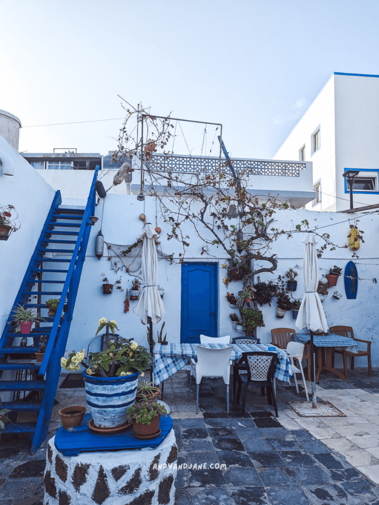 Charming courtyard in Corralejo, with blue stairs, potted plants, dining tables, and white walls adorned with greenery and colorful decor.