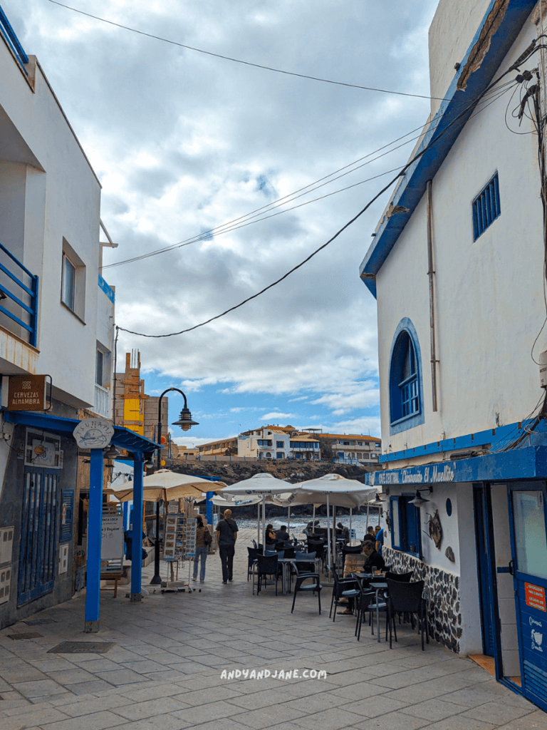 A charming coastal street in El Cotillo with white buildings and blue accents, featuring outdoor seating under umbrellas and a view of calm waters.