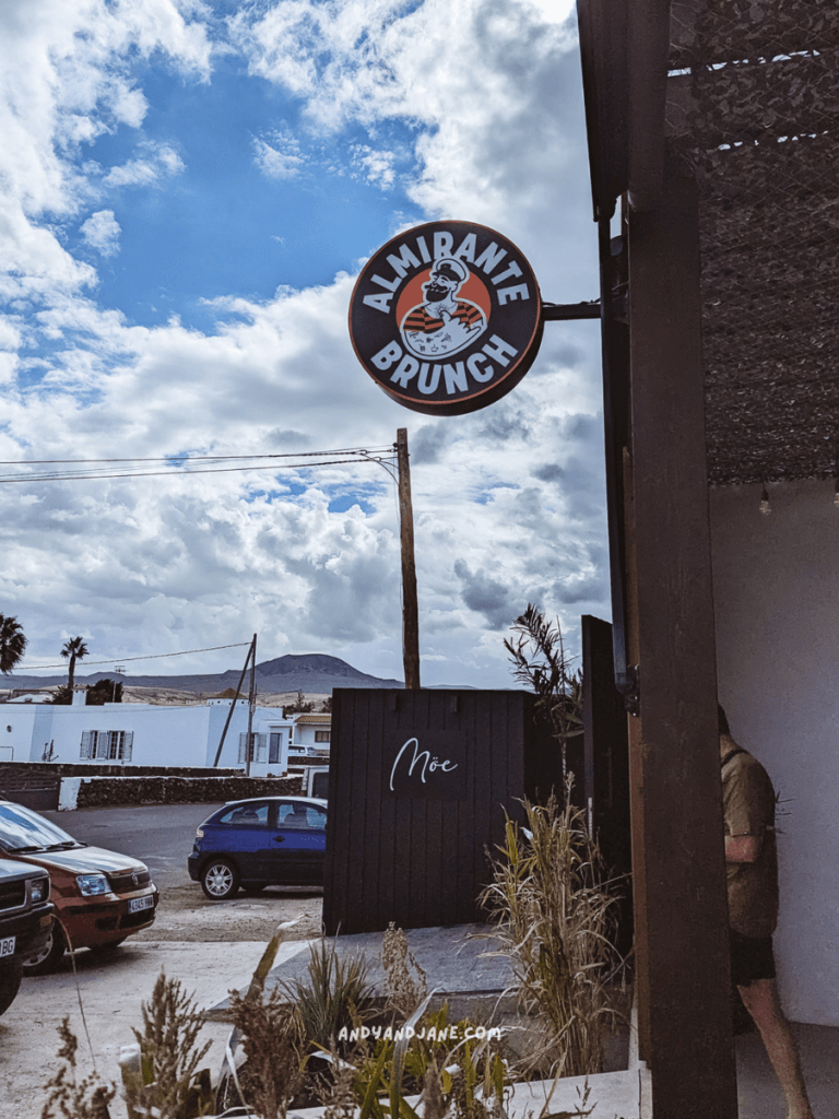 Sign for "Almirante Brunch" featuring a cartoon chef, set against a cloudy sky, with neighboring buildings and parked cars visible in Lajares.