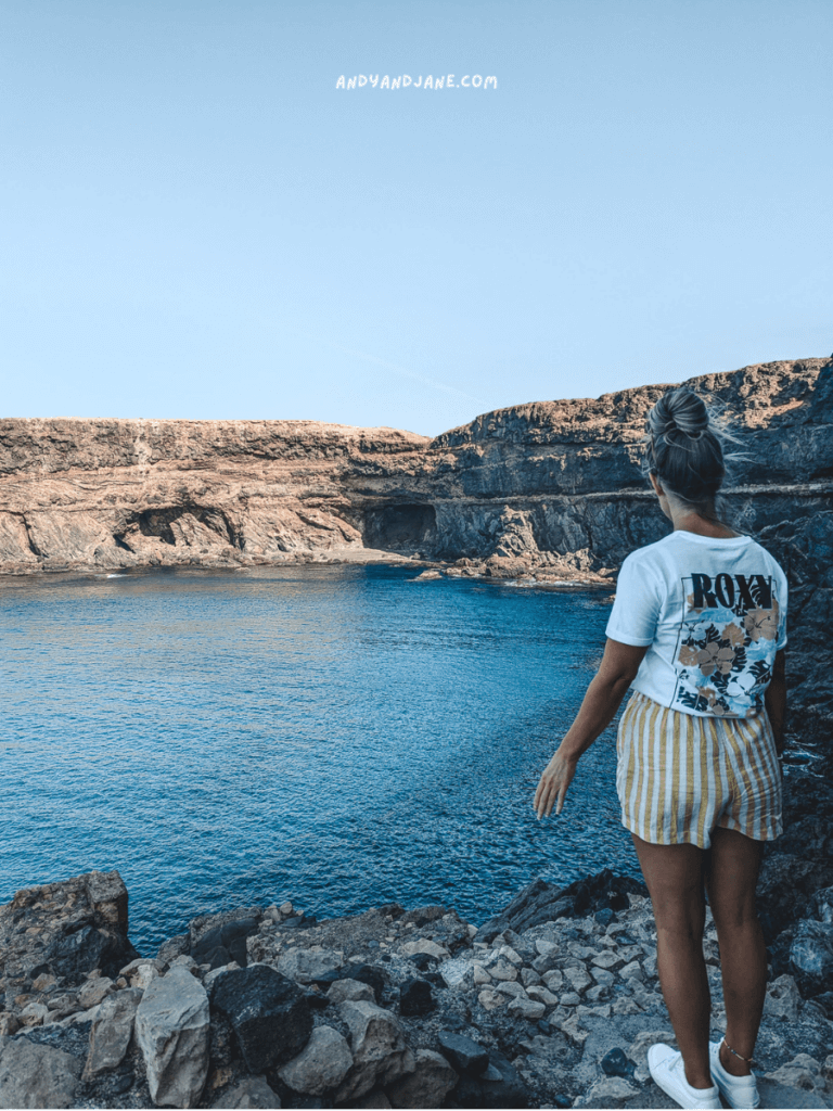 A woman in a Roxy t-shirt and striped shorts stands by a rocky coastline at the Ajuy Caves, gazing at a calm blue sea under a clear sky.