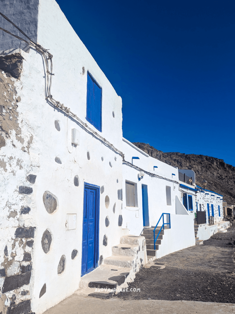 Whitewashed buildings in Pozo Negro with blue doors and windows, set against a clear blue sky and rocky landscape in a coastal village.