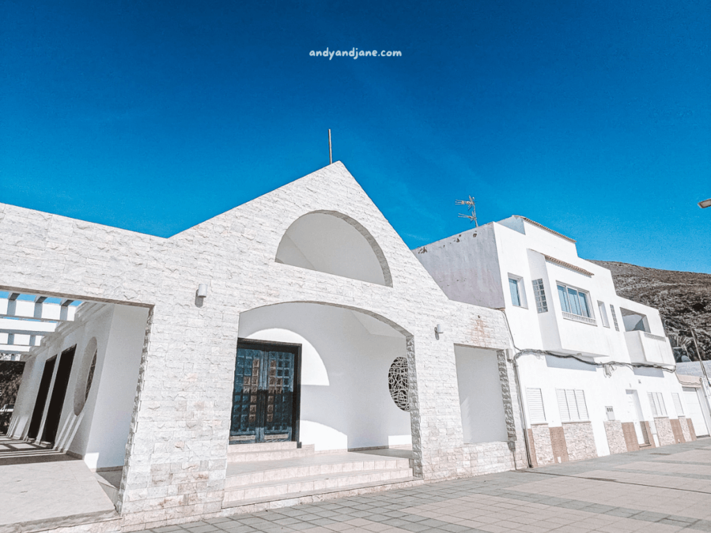 Modern white architecture featuring arched details and stone accents against a bright blue sky in Giniginamar Fuerteventura. Sunlit plaza with tiled ground.