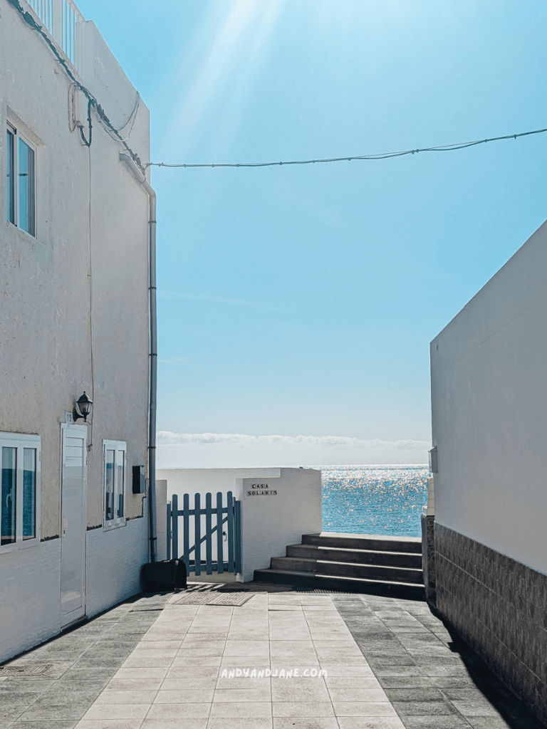 A sunlit pathway in Giniginamar between whitewashed buildings leads to a sparkling ocean view, framed by a blue gate and steps.