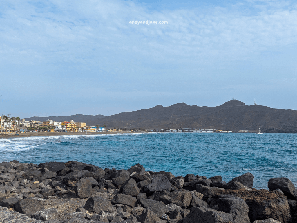 Coastal view of Gran Tarajal featuring rocky shoreline, blue waves, and a distant town with colorful buildings against a backdrop of hills and clouds.