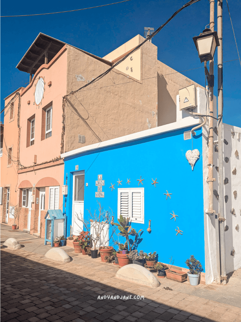 Colorful buildings in a quaint street in Giniginamar Fuerteventura, featuring a bright blue wall adorned with starfish and potted plants under a clear blue sky.