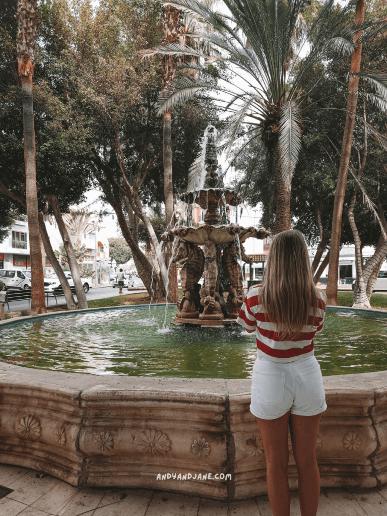 A woman in a red and white striped shirt stands by a decorative fountain surrounded by palm trees in a vibrant outdoor setting in Gran Tarajal, Fuerteventura.