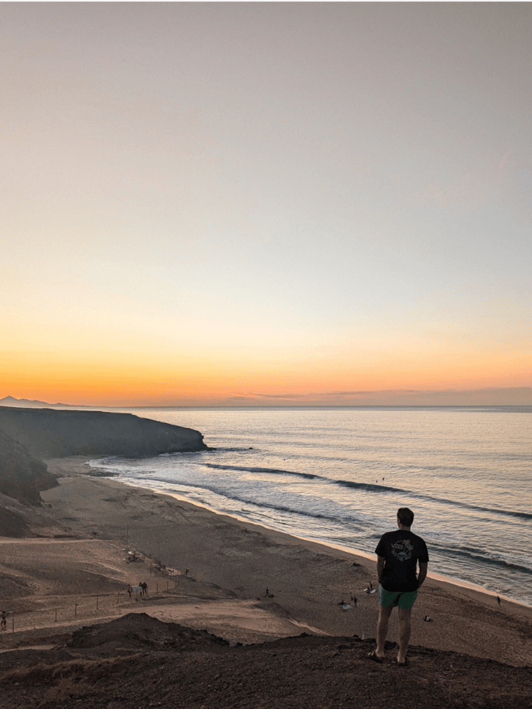 A person in a black shirt and green shorts stands on a cliff in La Pared Fuerteventura, overlooking a calm beach at sunset, with gentle waves lapping at the shore.