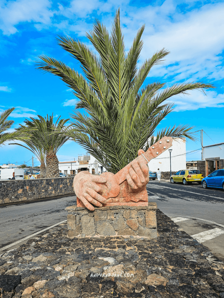 A vibrant sculpture of hands playing a guitar, surrounded by palm trees and a blue sky, along a lively street scene in Lajares, Fuerteventura.