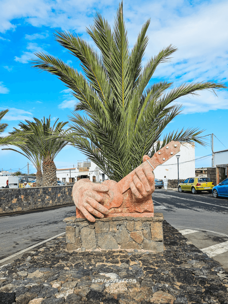 Stone sculpture of two hands playing a guitar, surrounded by palm trees and a blue sky in a vibrant town setting of Lajares, Fuerteventura.