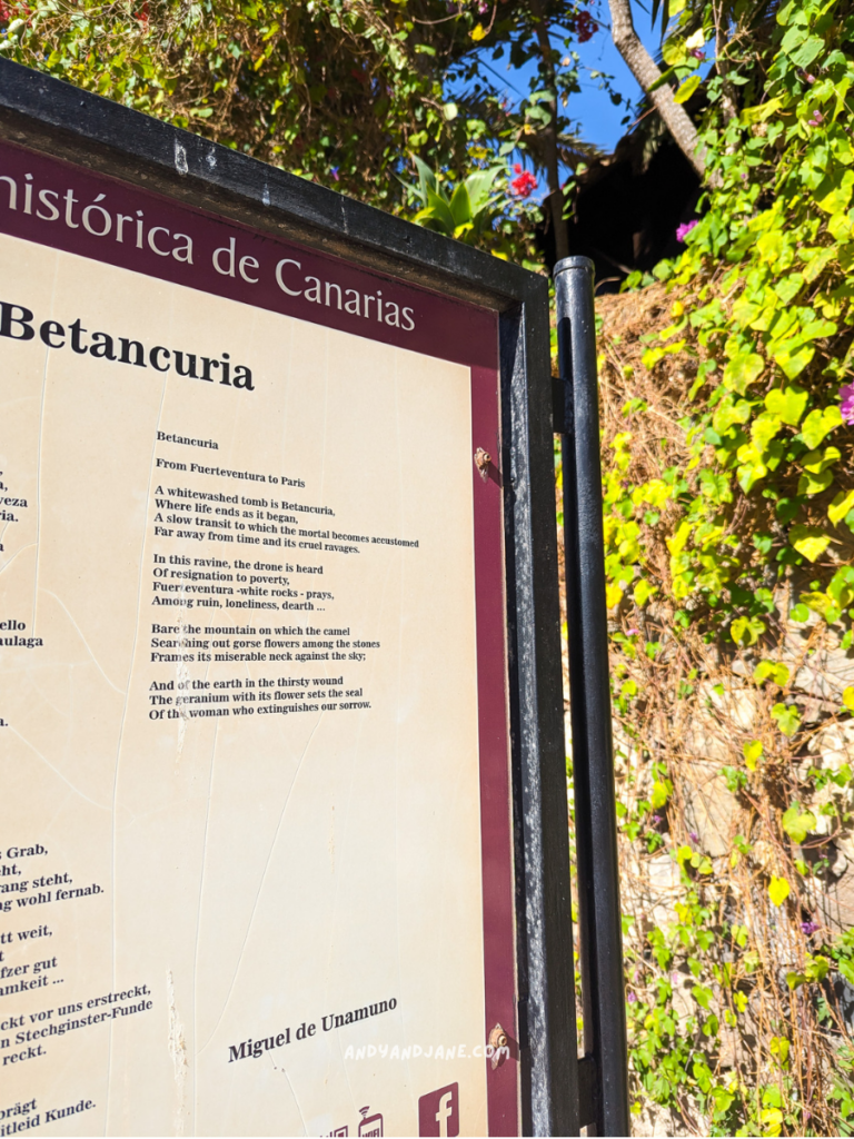 A sign for Betancuria in Fuerteventura, featuring text about its history and a poem by Miguel de Unamuno, surrounded by vibrant greenery.