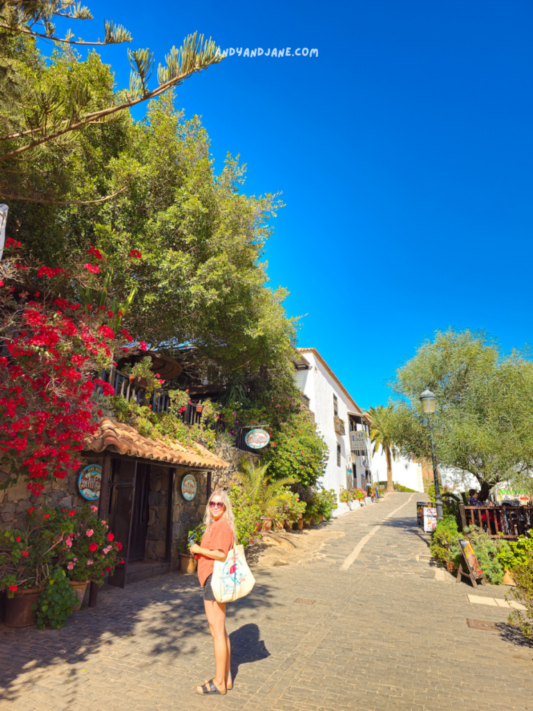 A sunny street scene in Betancuria featuring vibrant flowers, green trees, and charming white buildings, with a cobblestone path inviting exploration.