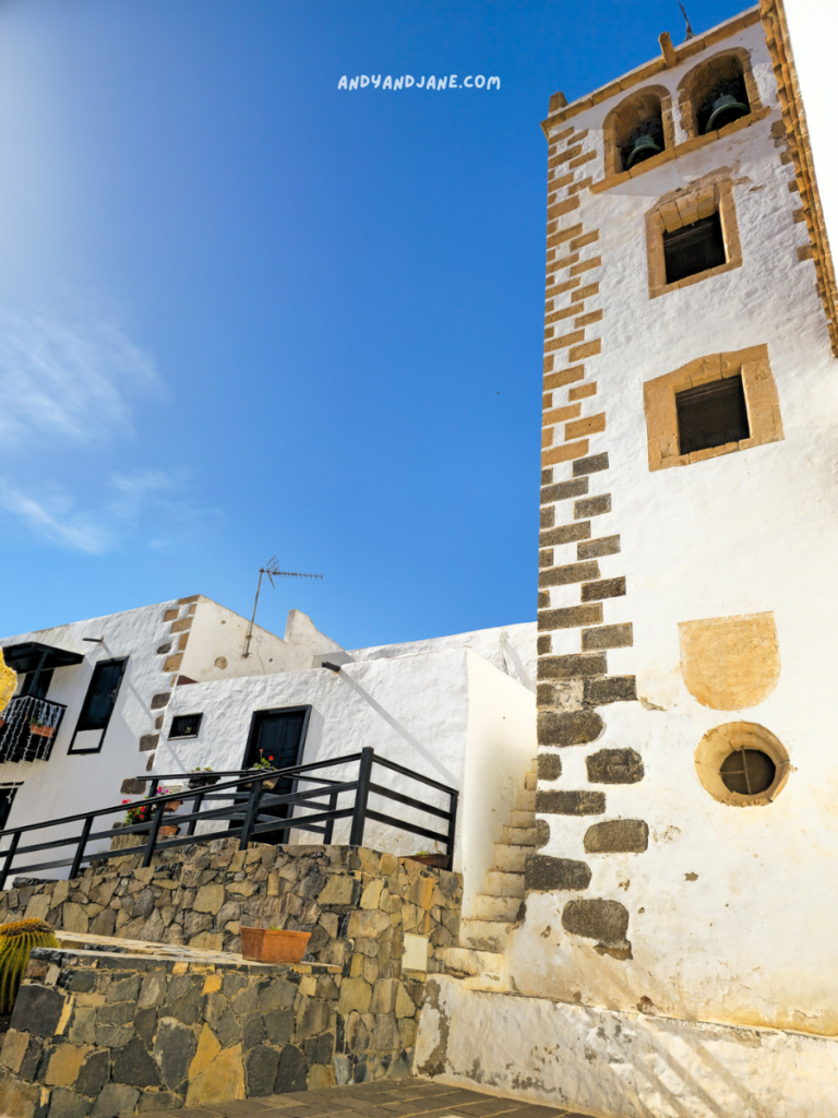 Charming whitewashed houses with black doors and balconies, surrounded by stone walls and colorful flowers, under a bright blue sky.