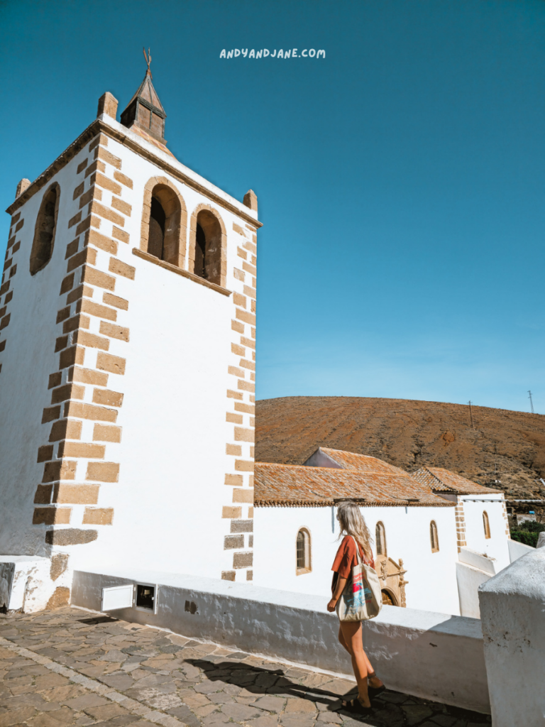 A woman stands near a historic white church with a bell tower, set against a clear blue sky and arid hills in the background.