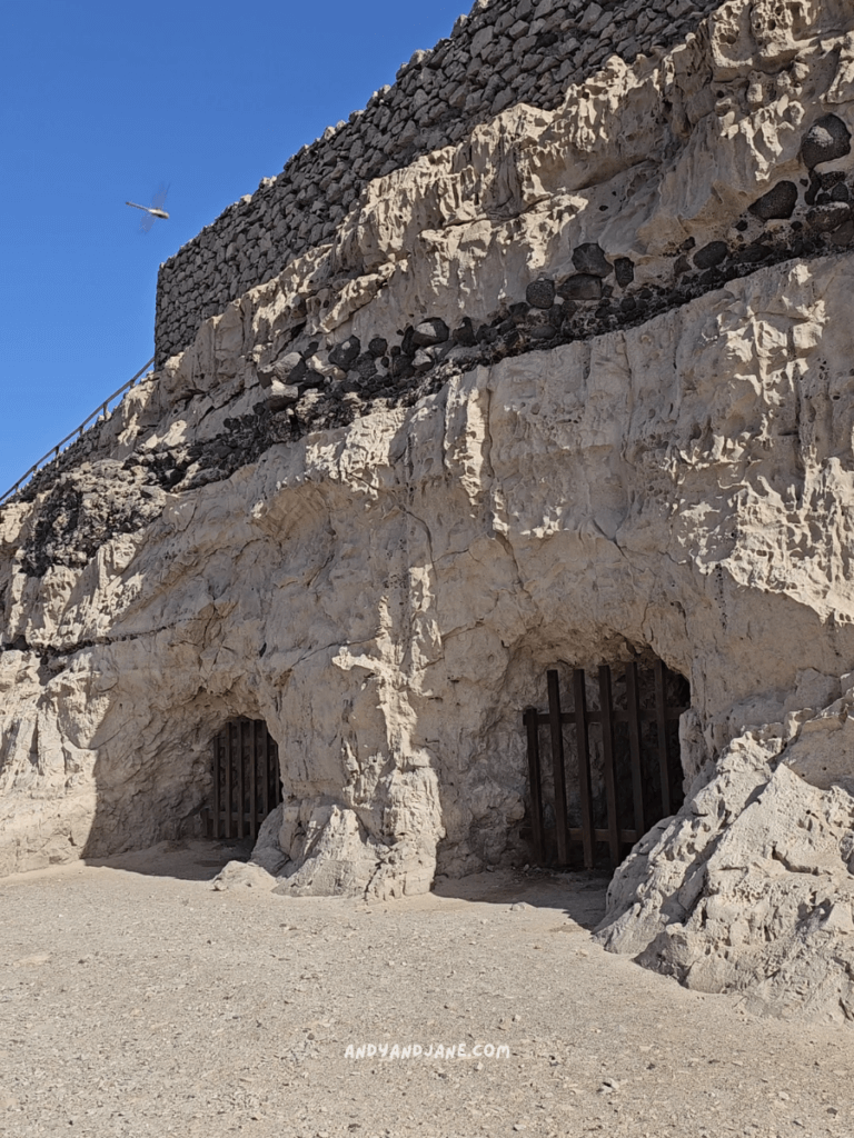 Two barred cave entrances at Ajuy in rocky terrain, with a stone wall above and a clear blue sky in the background.