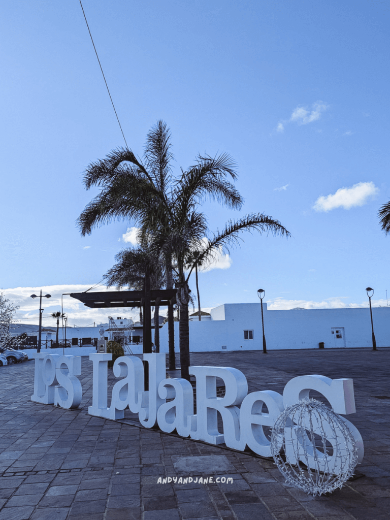 A sunny outdoor scene featuring large white letters spelling "Las Lajares," with palm trees and a clear blue sky in the background.