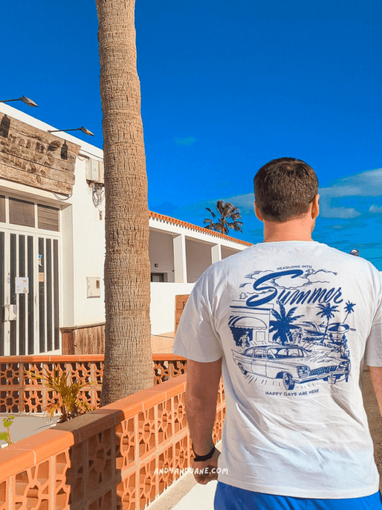 Person in a white t-shirt with a vintage car graphic, walking past a palm tree and sunny buildings, ready for summer.