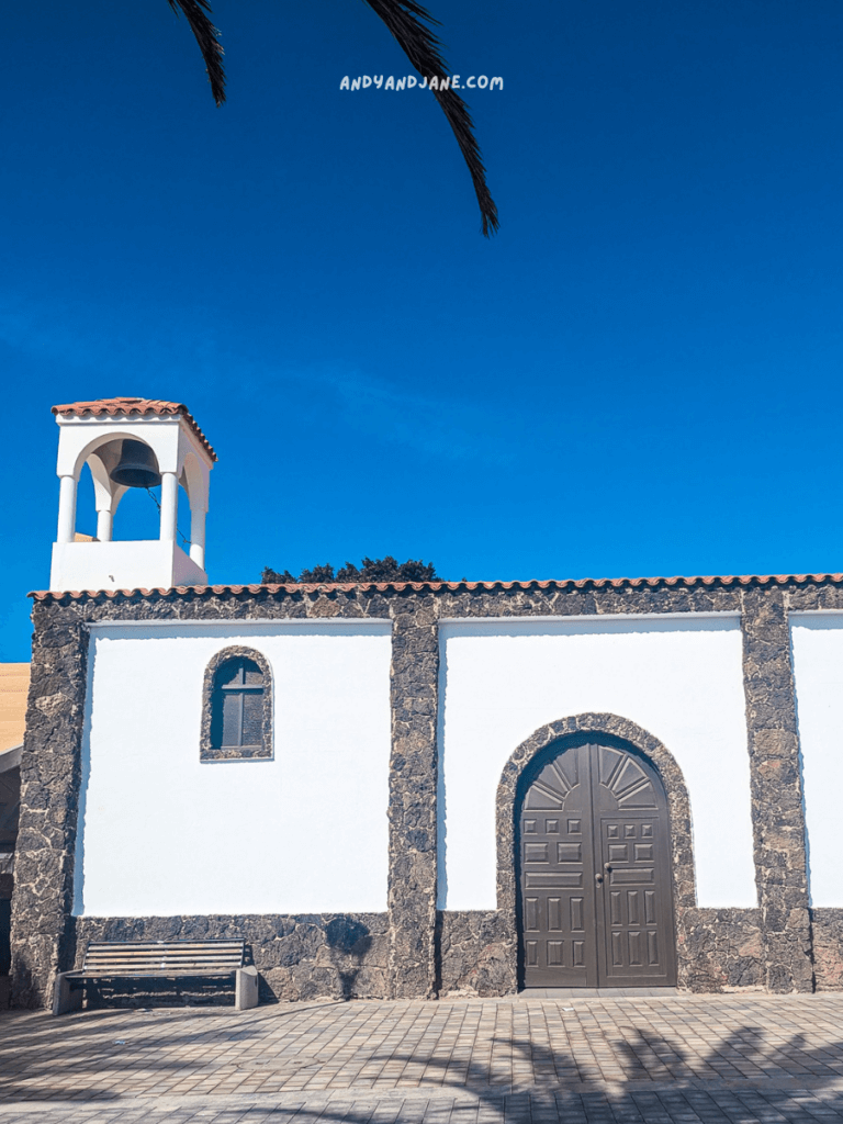 A whitewashed church in La Lajita with stone accents, featuring a bell tower and a double door, under a bright blue sky. Palm leaves partially shade the scene.