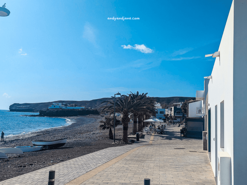 A scenic beachfront promenade in La Lajita, lined with palm trees and white buildings, featuring a pebbly beach and a calm bay under a blue sky.