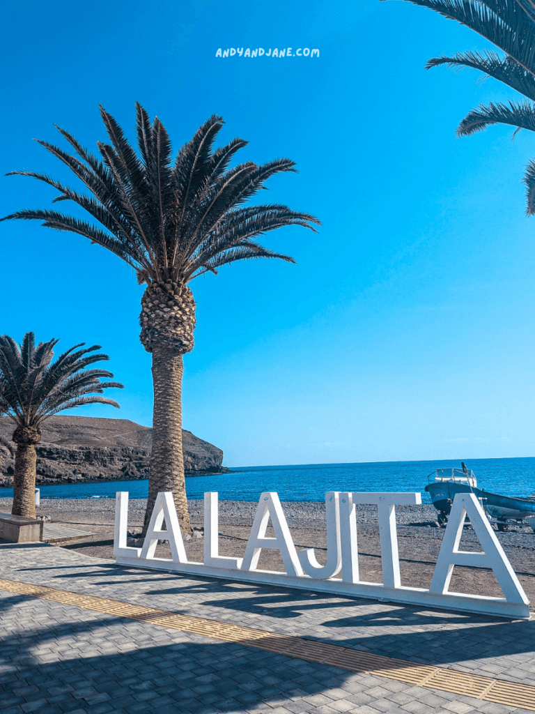 Beachfront sign reading "LA LAJITA" surrounded by palm trees, with a serene blue sky and ocean in the background. 