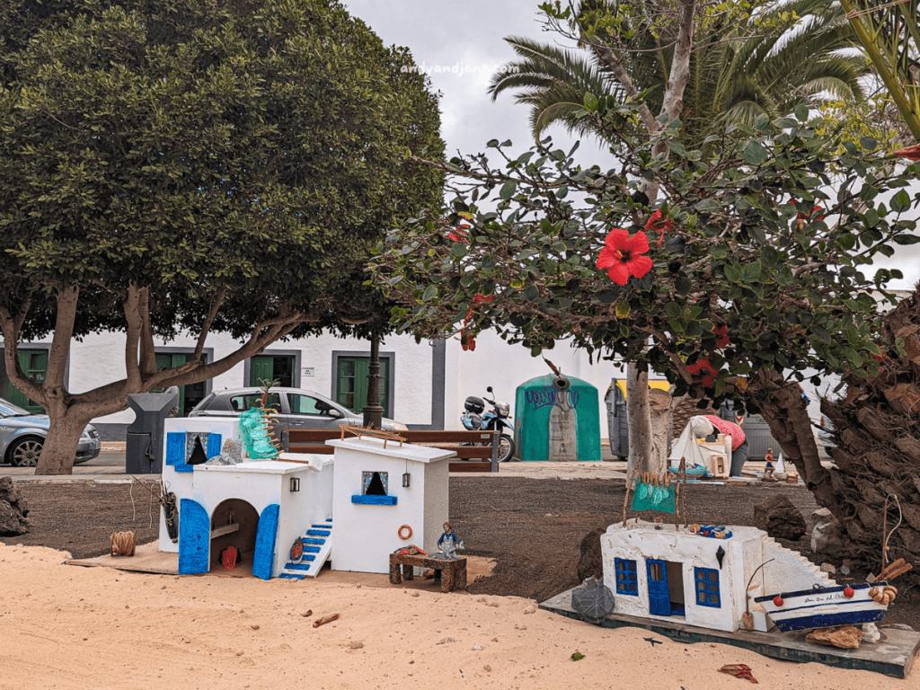 Colorful miniature houses in La Olvia, with blue accents and a big hibiscus flower, set against a sandy backdrop and palm trees.