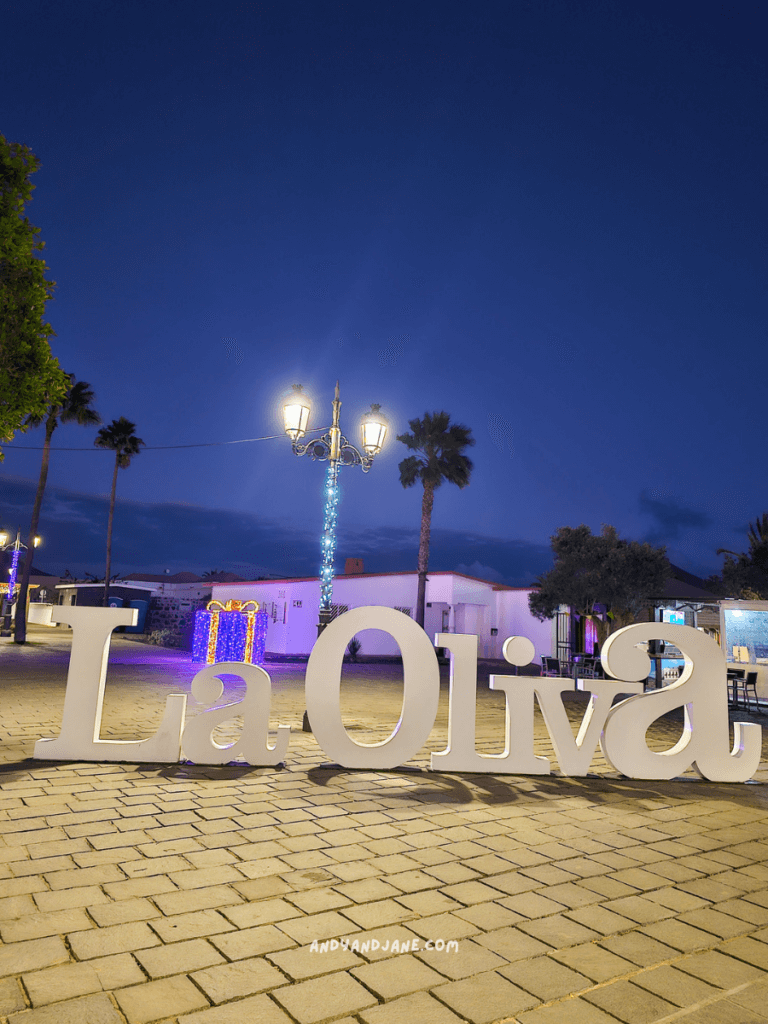 Illuminated sign reading "La Oliva" at dusk, surrounded by palm trees and street lamps, creating a vibrant atmosphere.