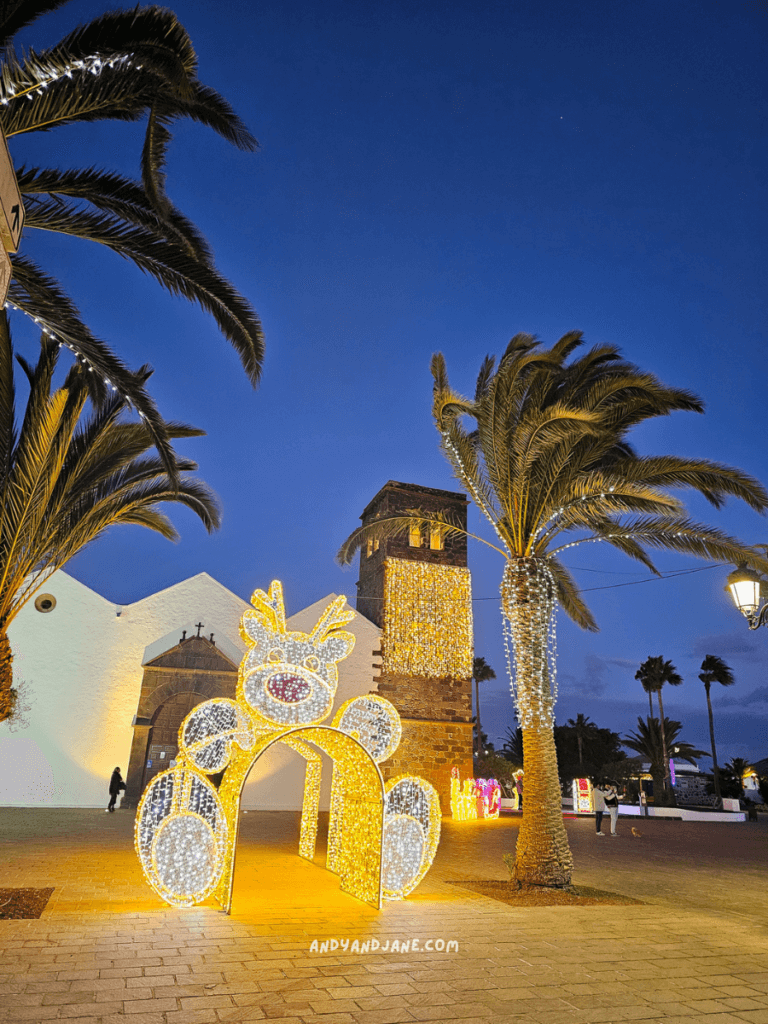 A glowing reindeer archway stands in front of a historic building, surrounded by palm trees, at dusk in La Oliva, Fuerteventura.