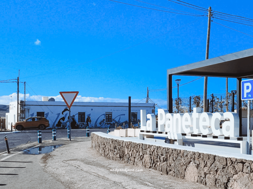 View of a street scene featuring a sign for "La Paneteca," a vibrant mural on a building, and a clear blue sky in Lajares.