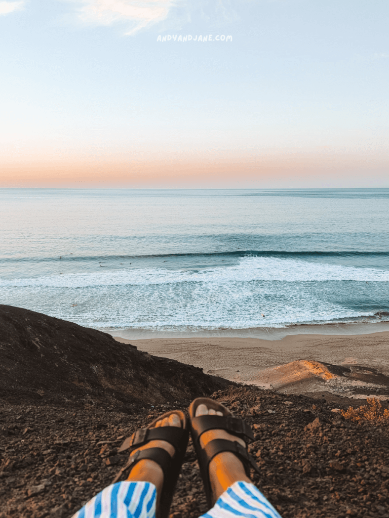 Bare feet in black sandals resting on rocky terrain, overlooking the tranquil beach at La Pared with gentle waves and a pastel sky at sunset.