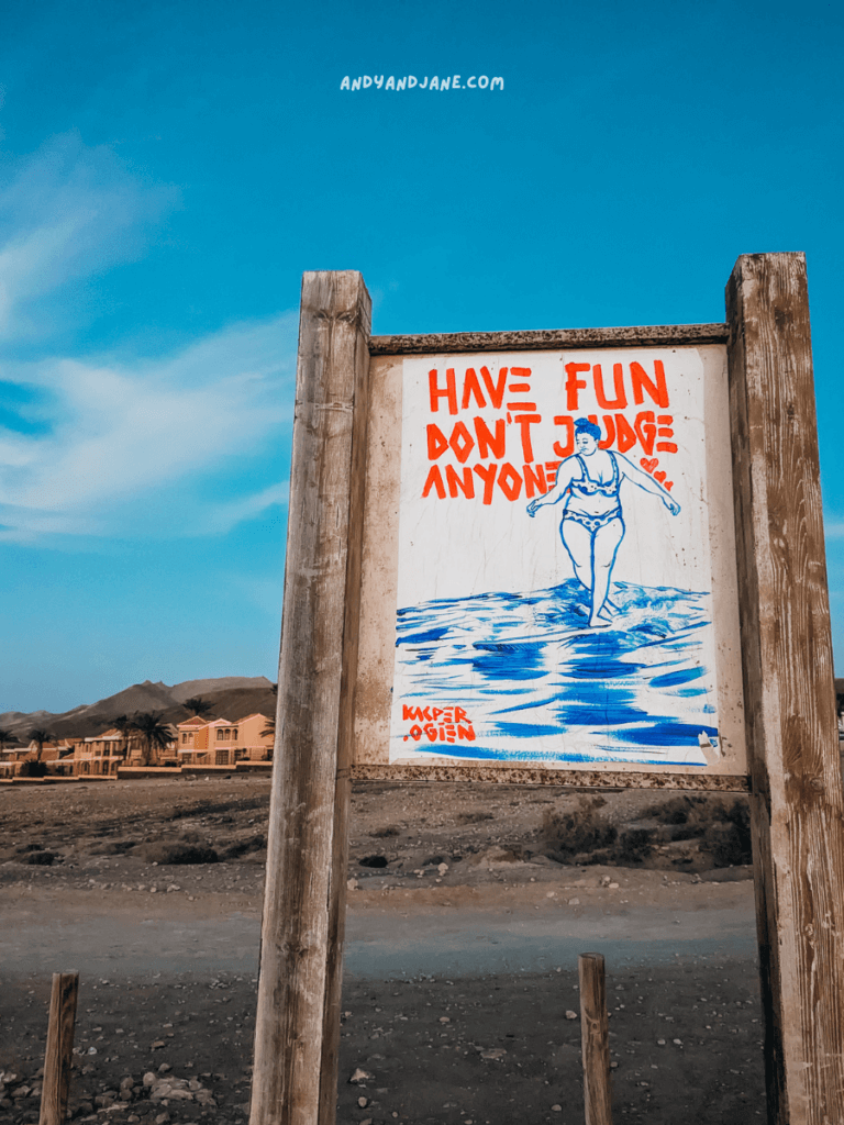 A colorful sign depicts a person walking in water, with the motto "Have fun, don't judge anyone" in bold letters. Scenic backdrop included.