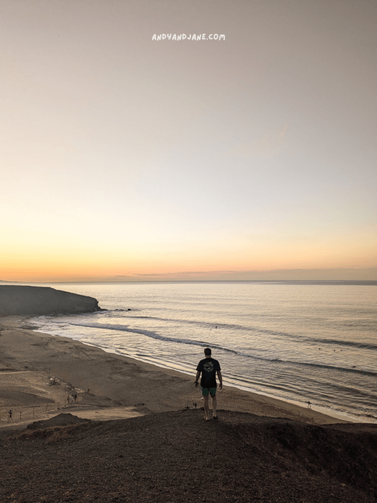 A man stands on a hill overlooking Viejo del Rey beach at sunset, with gentle waves lapping the shore and silhouettes of people in the distance.