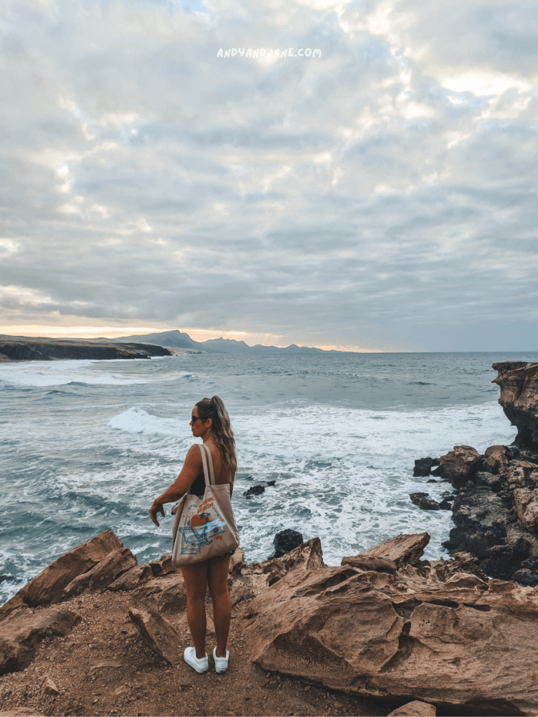 A person stands on rocky terrain by the sea in La Pared, gazing at the waves and distant mountains under a cloudy sky.