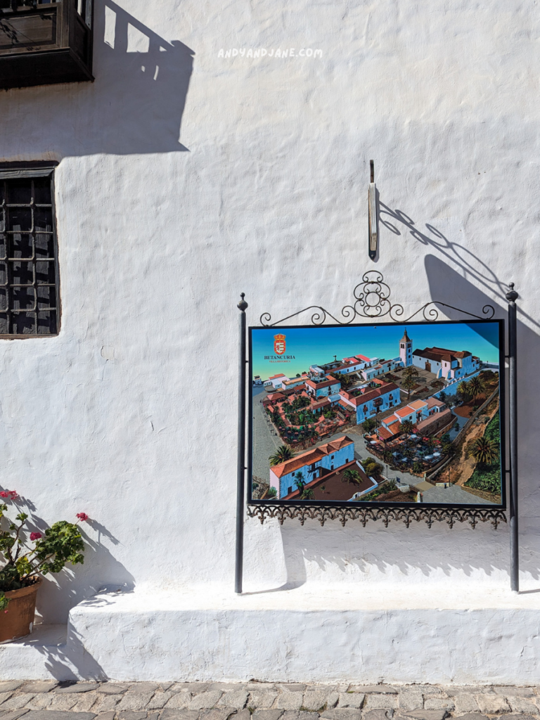 A colorful poster of Betancuria is displayed on a white wall, surrounded by a potted plant, with a cobblestone walkway below.