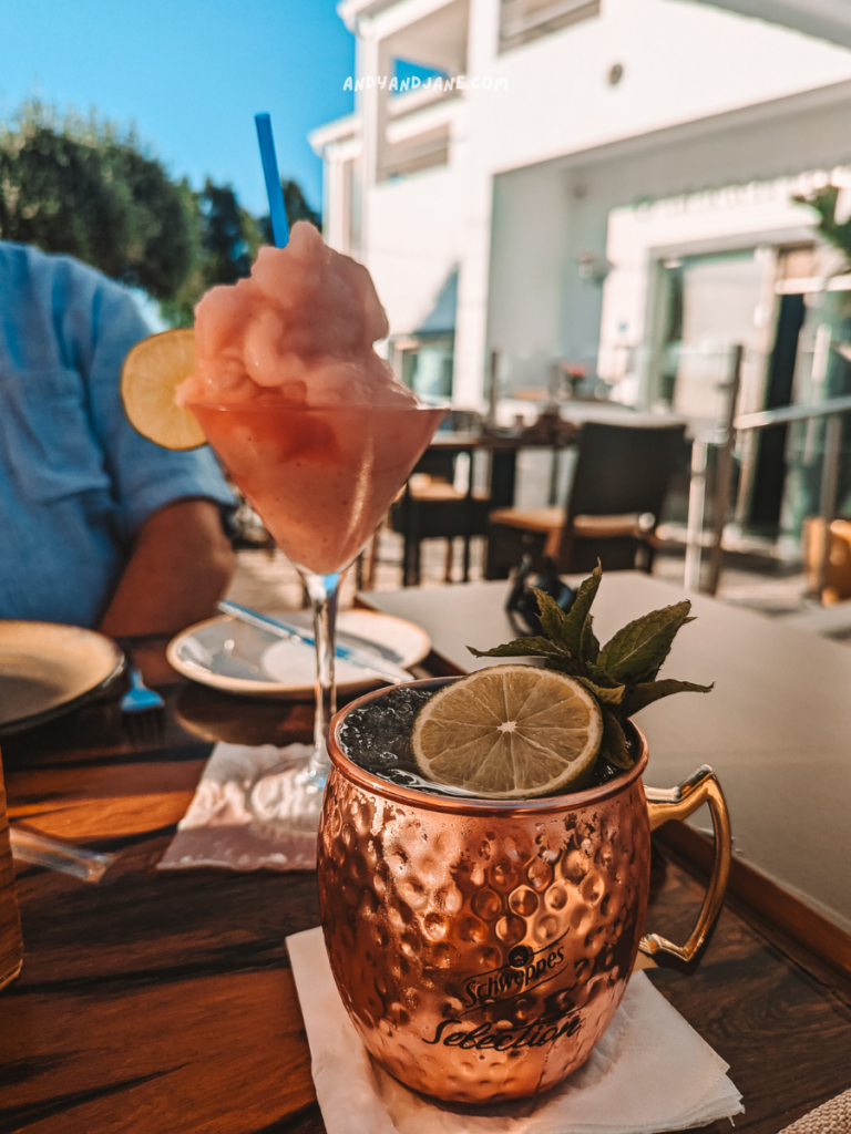 A colorful cocktail and slushy drink on a wooden table at Restaurante Marinheiro, Algarve, featuring a copper mug with mint and lime, set against a sunny backdrop.