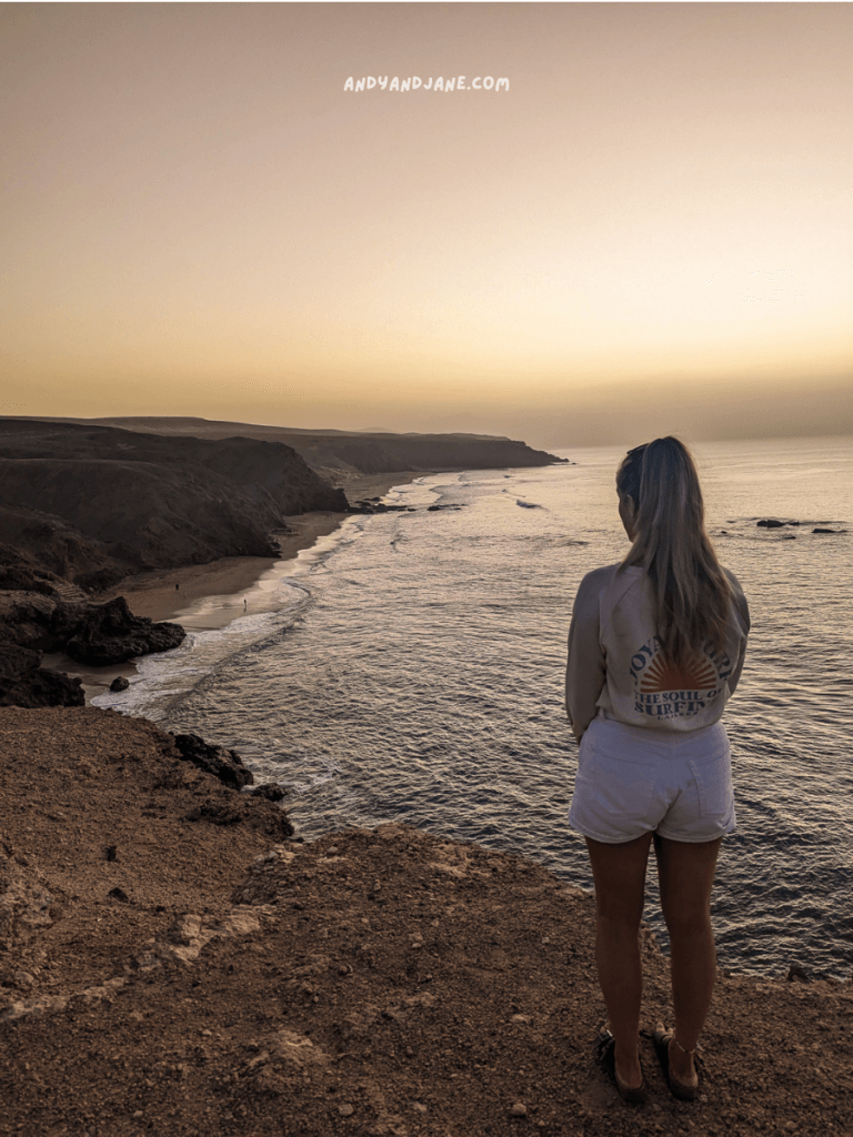 A woman stands on a rocky cliff in La Pared, Fuerteventura gazing at the serene beach and ocean during sunset, with soft pastel skies in the background.