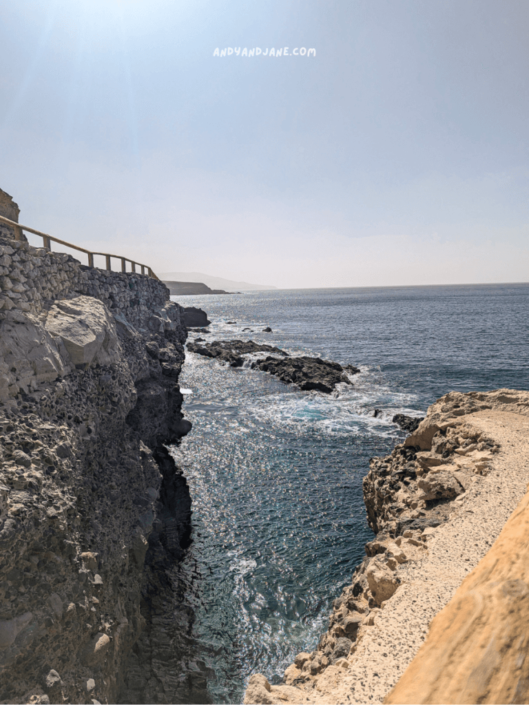 A coastal view on the Ajuy Caves walk showcasing rocky cliffs, a wooden railing, and serene blue waters under a clear sky.