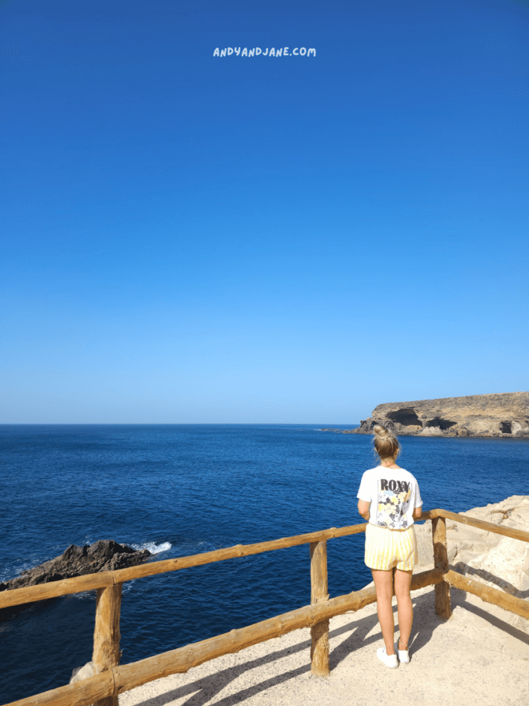 A woman in a Roxy t-shirt and yellow shorts stands on a coastal lookout, gazing at the serene blue ocean under a clear sky in Ajuy, Fuerteventura