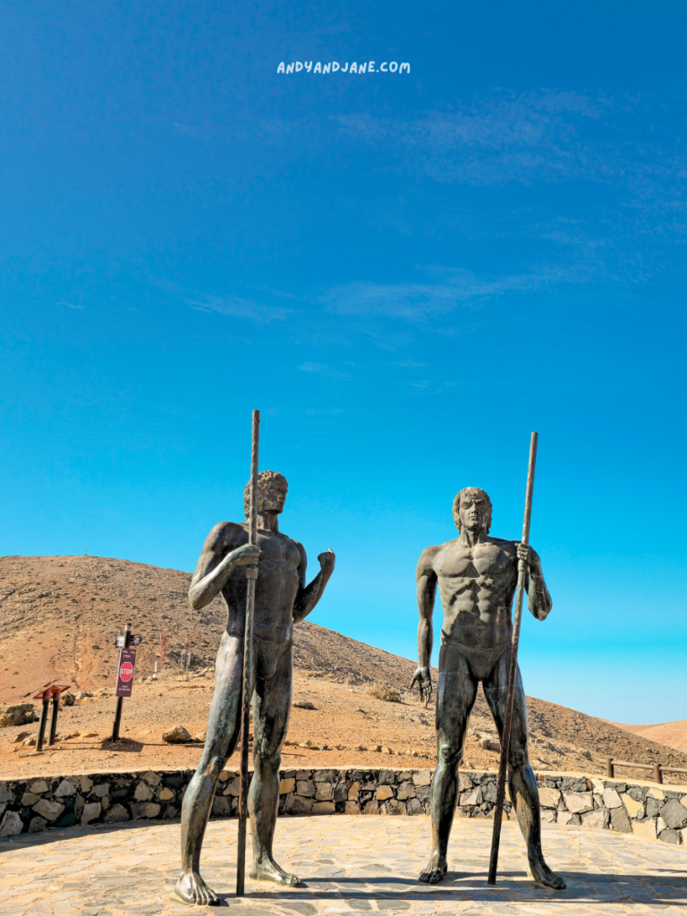 Two life-sized bronze statues of muscular figures stand on a stone platform, each holding a long pole. The figure on the left has a slight smile and appears to be in a relaxed pose, while the figure on the right stands confidently with a serious expression. In the background, a dry, mountainous landscape extends under a clear blue sky. The scene conveys a sense of strength and resilience, highlighting the artistic depiction of these statues.