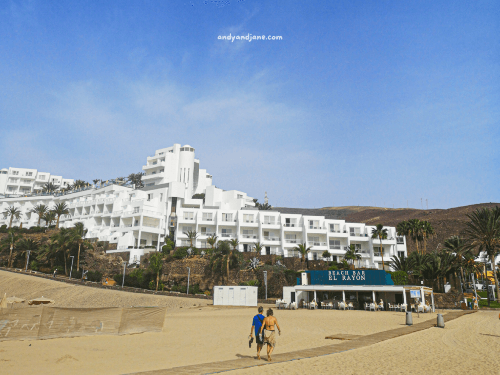 A white hotel overlooks the beach in Morro Jable, with palm trees lining the sandy shore and a beach bar named "El Rayón" in the foreground.