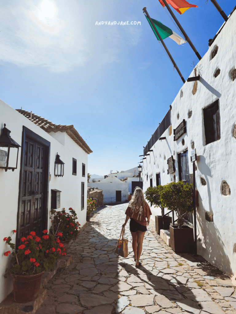 A woman with long hair walks down a cobblestone street in Betancuria, Fuerteventura, lined with white buildings and vibrant red flowers under a clear blue sky.