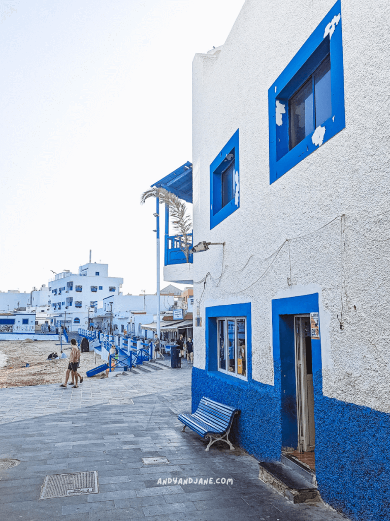 A charming seaside walkway in Corralejo featuring white buildings with blue accents, a bench, and people strolling near the beach.