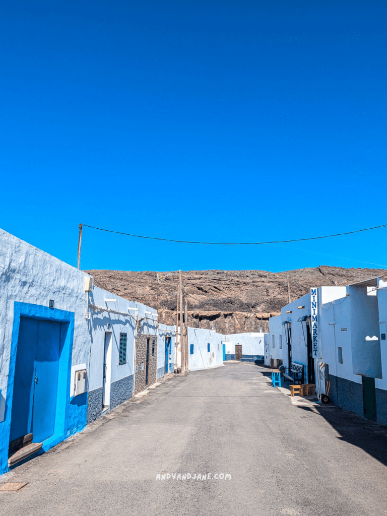 A narrow street lined with white and blue houses under a clear blue sky, with a mini market on the right. Rocky hills in the background in Ajuy, Fuerteventura.