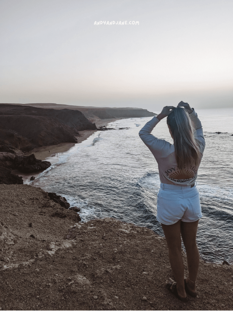 A woman with long hair stands on a cliff, gazing at the serene ocean and coastline during dusk in La Pared, Fuerteventura.