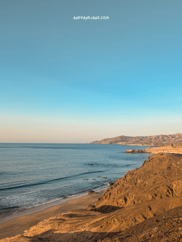 A serene coastal view with gentle waves lapping against sandy shores, framed by rocky cliffs under a clear blue sky.