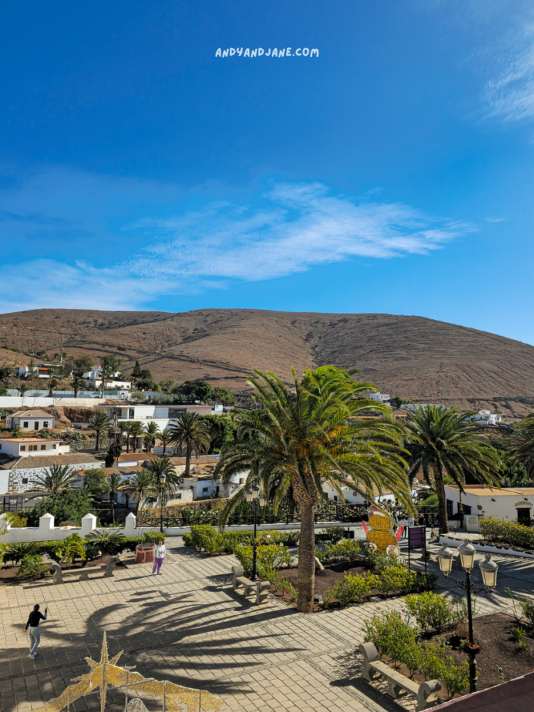 Two people stand in a sunlit plaza surrounded by palm trees, overlooking scenic hills and white buildings under a blue sky.