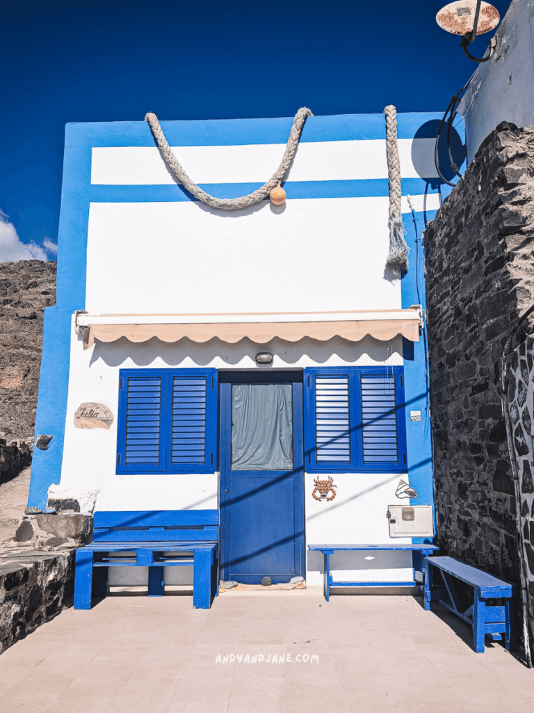 Charming white building in Pozo Negro with blue accents, featuring wooden benches and decorative elements, set against a bright blue sky.