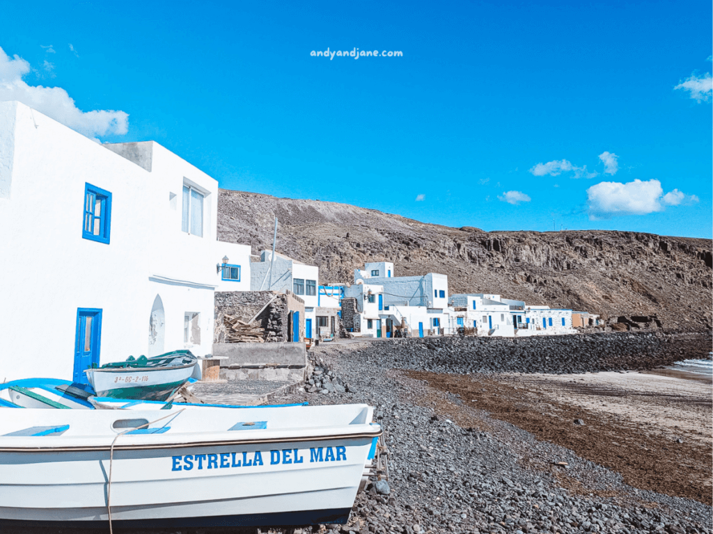 A white fishing boat named "Estrella del Mar" on a rocky shore, with coastal white houses and a clear blue sky in the background in Pozo Negro, Fuerteventura.