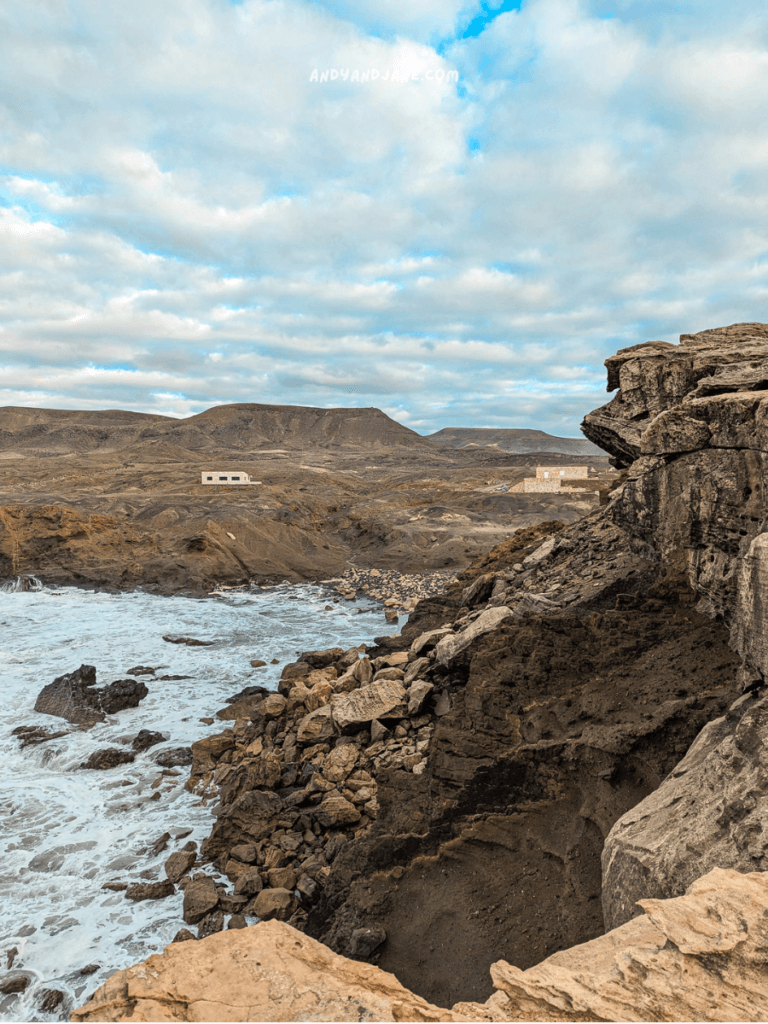 Rocky coastline at La Pared, Fuerteventura with crashing waves, distant mountains, and a solitary white building under a cloudy sky.